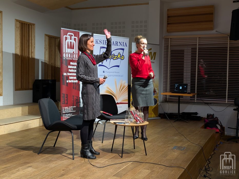 Biblioteka Gorlice, „Mój przyjaciel Masaj” — spotkanie autorskie z Elżbietą Wiejaczką na scenie stoi z uniesioną ręką w geście powitania autorka, Elżbieta Wiejaczka i z mikrofonem rozpoczynając spotkanie moderatorka, Joanna Kalisz-Dziki