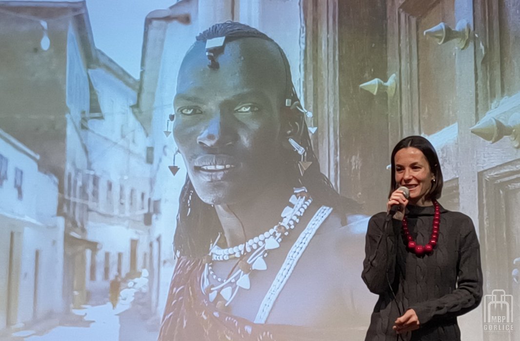 Biblioteka Gorlice, „Mój przyjaciel Masaj” — spotkanie autorskie z Elżbietą Wiejaczką Elżbieta Wiejaczka stoi na tle dużego zdjęcia z Isają - Masajem i mówi do mikrofonu