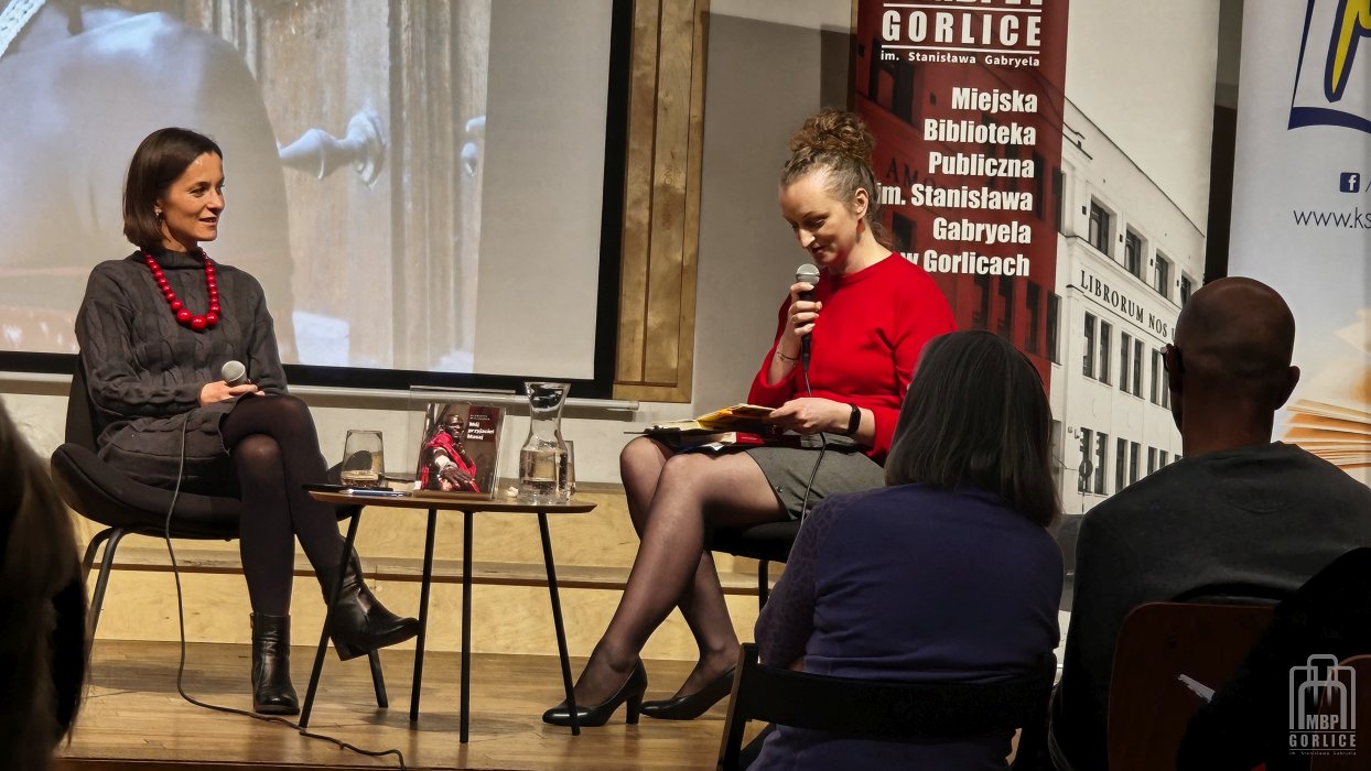 Biblioteka Gorlice, „Mój przyjaciel Masaj” — spotkanie autorskie z Elżbietą Wiejaczką bliski kadr na autorkę, która słucha i moderatorkę, która czyta fragment z książki