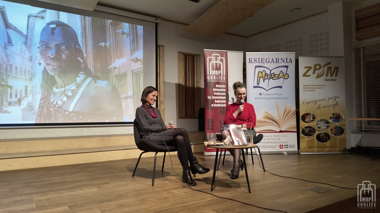 Biblioteka Gorlice, „Mój przyjaciel Masaj” — spotkanie autorskie z Elżbietą Wiejaczką na scenie siedzi uśmiechnięta autorka a moderatorka z mikrofonem zwraca się do publiczności