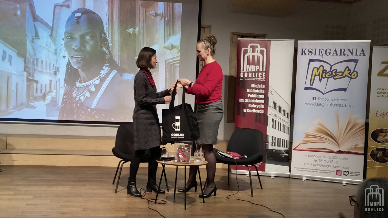 Biblioteka Gorlice, „Mój przyjaciel Masaj” — spotkanie autorskie z Elżbietą Wiejaczką na scenie stoją Elżbieta Wiejaczka i Joanna Kalisz-Dziki, która dziękuje i wręcza pamiątkowe torby