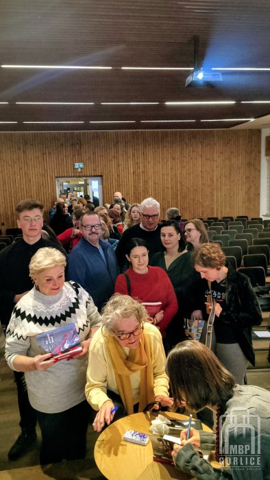 Biblioteka Gorlice, „Mój przyjaciel Masaj” — spotkanie autorskie z Elżbietą Wiejaczką autorka wpisuje dedykację do książki, przed nią kolejka osób przez całą salę
