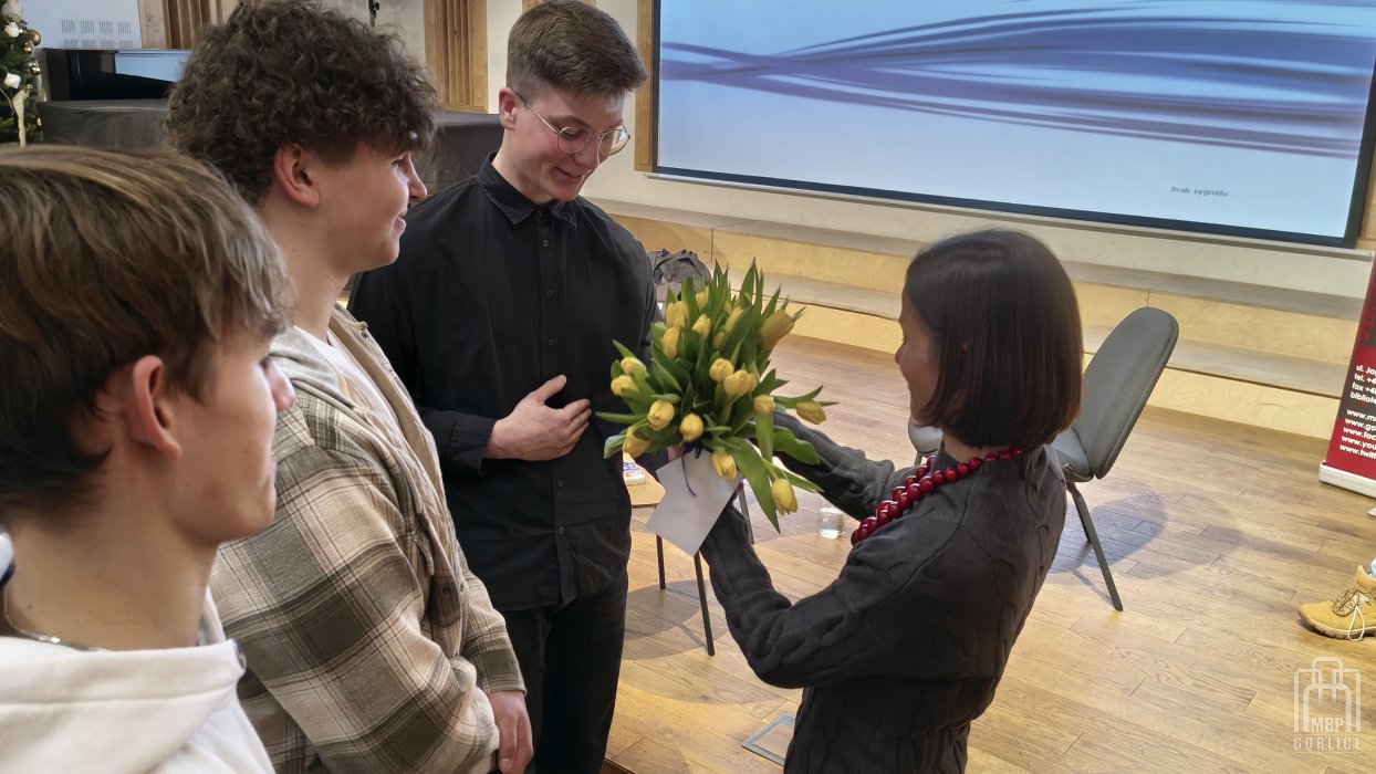 Biblioteka Gorlice, „Mój przyjaciel Masaj” — spotkanie autorskie z Elżbietą Wiejaczką trzech uczniów Liceum Ekonomicznego w Gorlicach wręcza autorce bukiet tulipanów