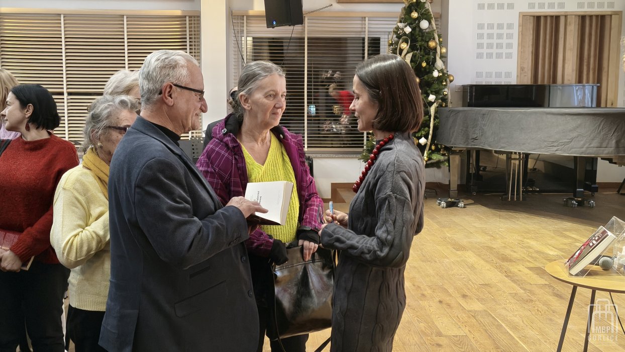 Biblioteka Gorlice, „Mój przyjaciel Masaj” — spotkanie autorskie z Elżbietą Wiejaczką autorka stoi obok sceny i rozmawia z uczestniczką i uczestnikiem spotkania