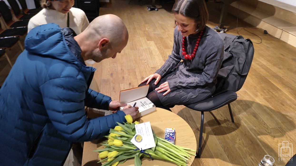 Biblioteka Gorlice, „Mój przyjaciel Masaj” — spotkanie autorskie z Elżbietą Wiejaczką autorka siedzi na scenie uśmiechnięta jej mąż pochylony nad stolikiem składa autograf w książce