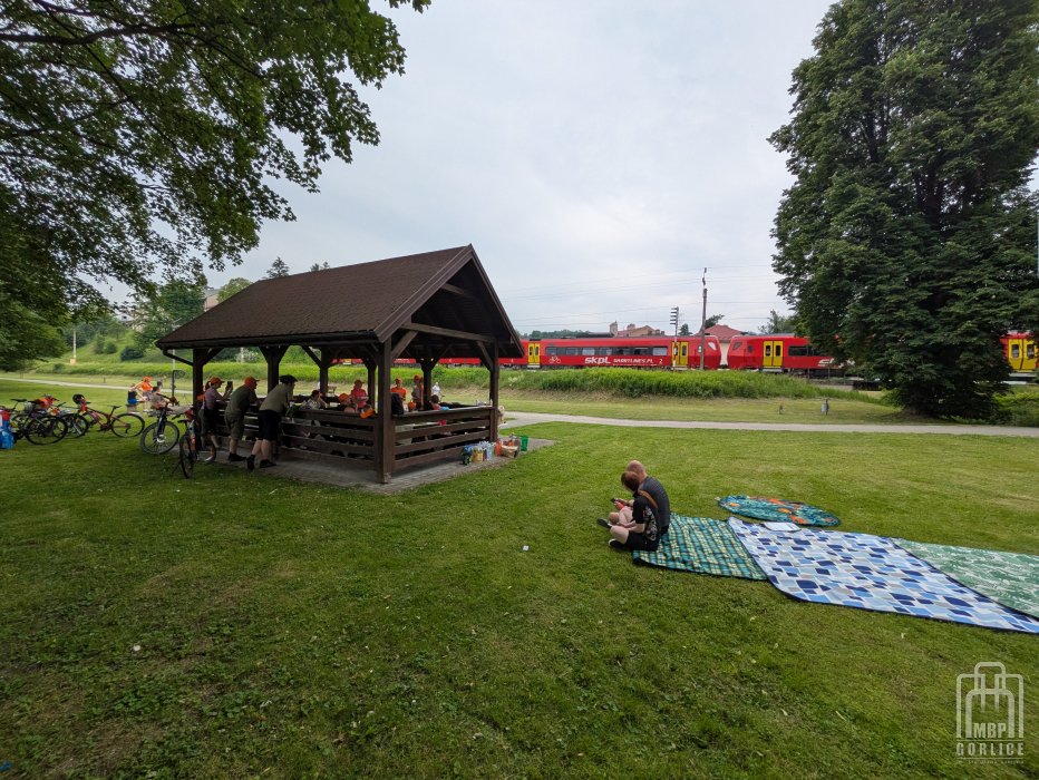 Biblioteka Gorlice, Odjazdowy Bibliotekarz zdjęcie z oddali na wiatę z uczestnkami po lewej widac zaparkwoane rowery po prawej dwie osoby siedzą na koach a w tle jedzie pociąg