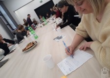 Powiększ zdjęcie Uśmiechnięta%20bibliotekarka%20siedzi%20przy%20stole%20i%20pisze%20na%20kartce,%20w%20tle%20inni%20uczestnicy%20szkolenia